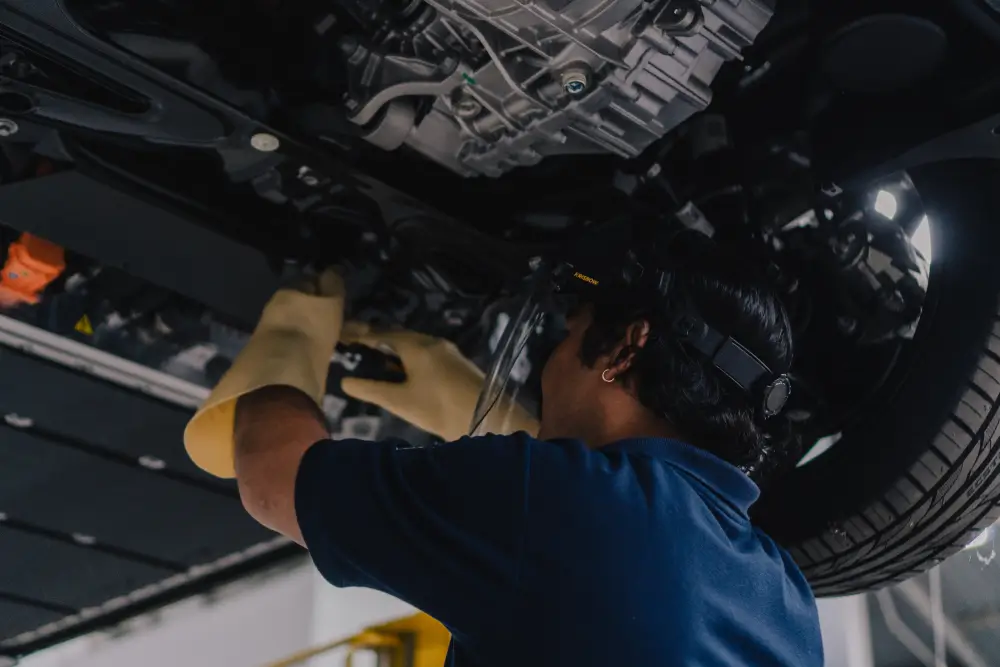 Teknisi VinFast dengan sarung tangan kuning dan pelindung wajah sedang memeriksa paket baterai di bagian bawah mobil listrik yang terangkat di bengkel.