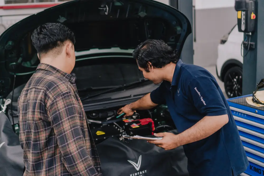 Seorang teknisi VinFast berseragam biru menunjuk ke bagian mesin mobil listrik, menjelaskan kepada seorang pelanggan laki-laki yang mendengarkan.