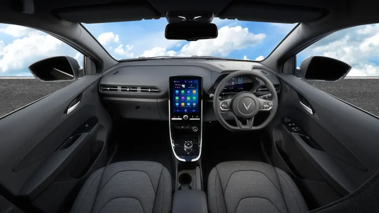 Tampilan interior kabin depan mobil listrik VinFast VFe34, terlihat dari kursi belakang. Latar belakang jalan raya dan langit biru berawan terlihat melalui kaca depan.
