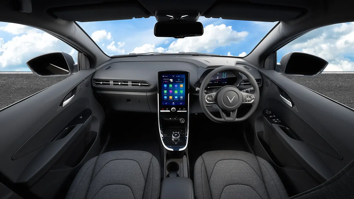 Tampilan interior kabin depan mobil listrik VinFast VFe34, terlihat dari kursi belakang. Latar belakang jalan raya dan langit biru berawan terlihat melalui kaca depan.